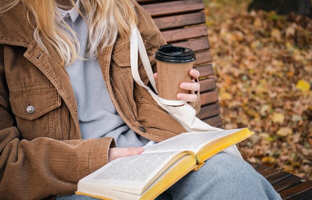 Молодая женщина сидит на скамейке в осеннем парке, пьет кофе и читает книгу