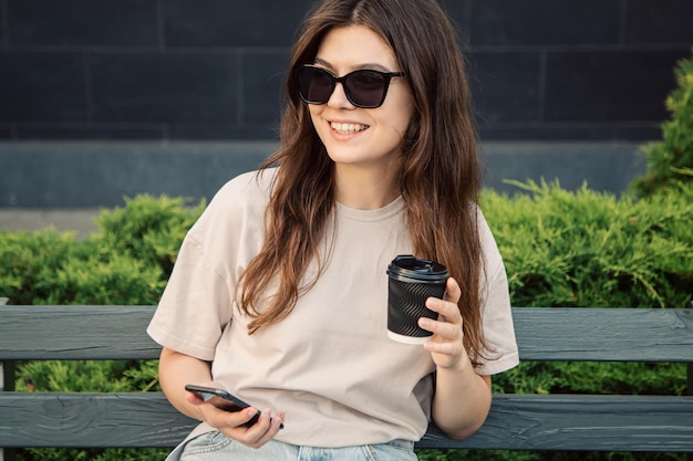 A young woman sits on a bench with a cup of coffee and a smartphone