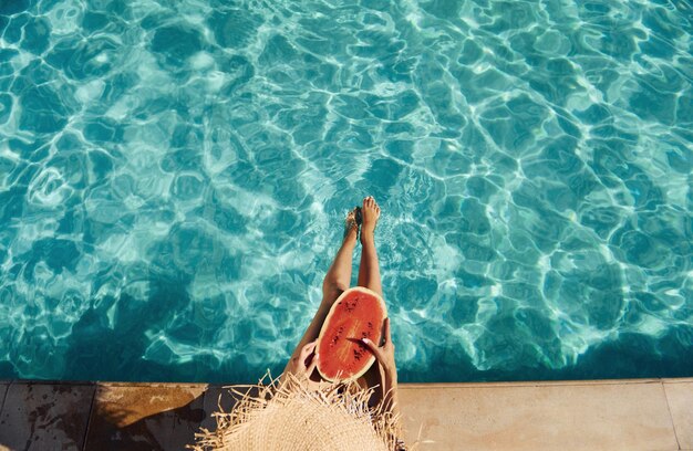 Young woman sits near swimming pool at daytime with watermelon