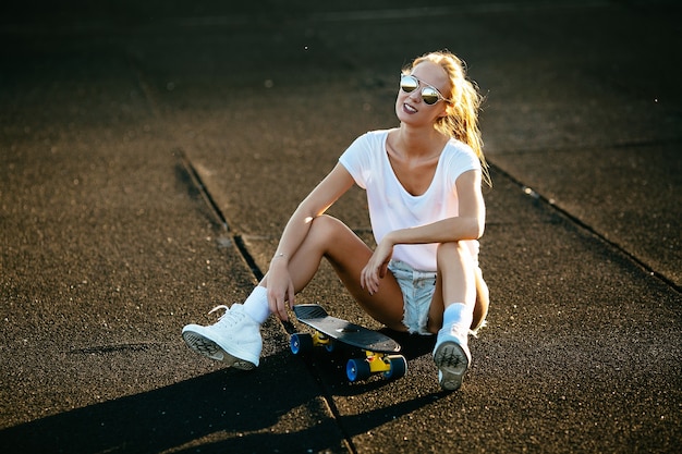 Бесплатное фото Молодая женщина сидит на скейтборде с солнцезащитными очками во время заката.
