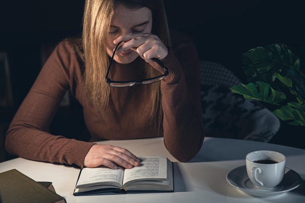 Молодая женщина сидит за столом и читает книгу ночью при свете лампы