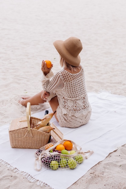 A young woman sits on the towel in a straw hat and a white knitted clothes with picnic basket