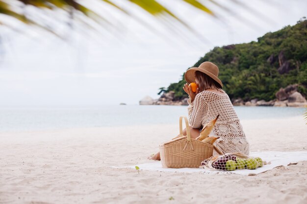 A young woman sits on the towel in a straw hat and a white knitted clothes with picnic basket