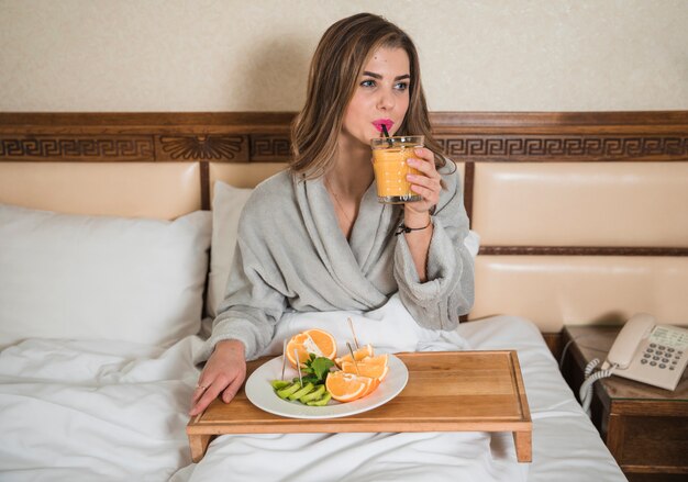 ジュースのガラスを飲むベッドの上に座っている若い女性