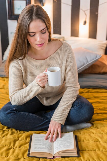 Young woman sitting on bed holding cup of coffee reading book