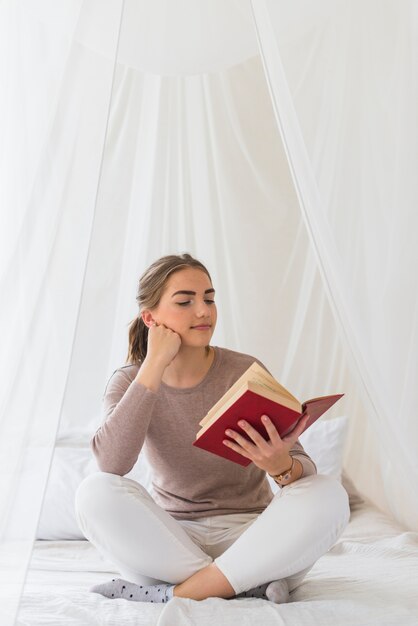 Young woman sitting on bed reading book