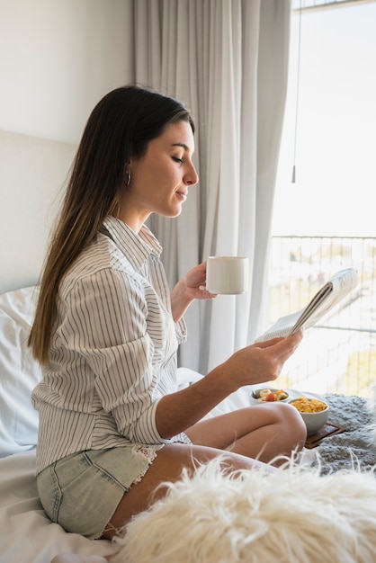 Young woman sitting on bed reading newspaper while drinking coffee