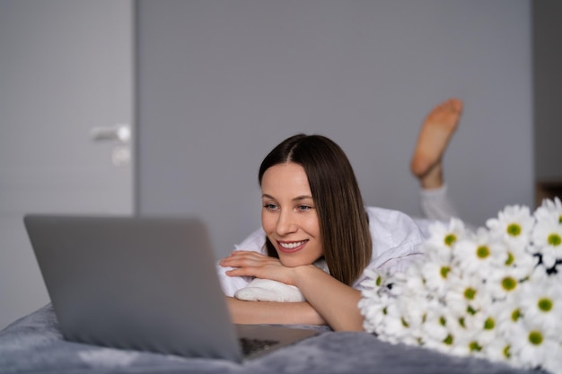 Молодая женщина, сидящая на кровати в пижаме, с удовольствием наслаждается белыми цветами и болтает с помощью ноутбука