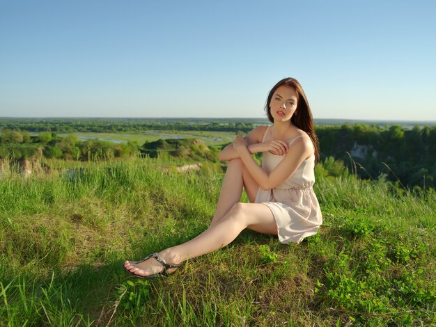 Молодая женщина, сидя у скалы на открытом воздухе на природе. Привлекательная девушка в белом платье позирует на открытом воздухе. Женская модель позирует в поле в солнечный летний день.