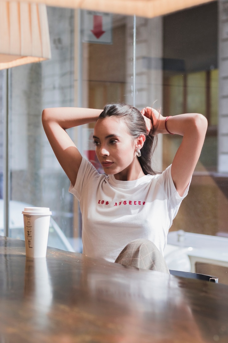 無料Photo テーブルの上の使い捨てのコーヒーカップと彼女の髪を結ぶカフェに座っていた若い女性