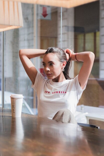 Young woman sitting in the caf� tying her hair with disposable coffee cup on table