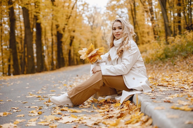 Молодая женщина сидит на обочине в осеннем лесу. Блондинка держит желтые листья. Девушка в бежевом пальто и коричневых брюках.