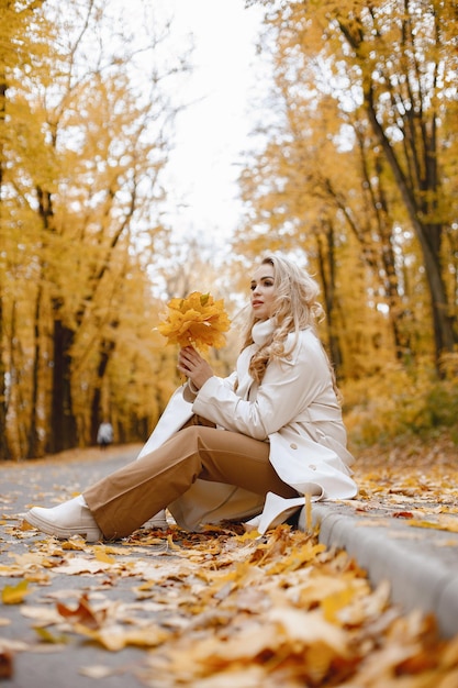 Young woman sitting on a curb in autumn forest. Blonde woman holding a yellow leaves. Girl wearing beige coat and brown trousers.