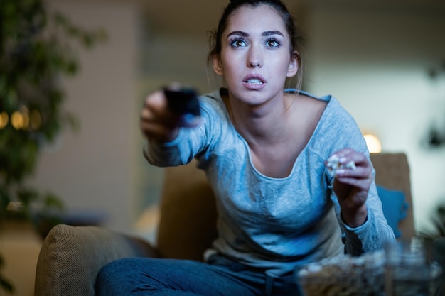 Giovane donna seduta al buio e che cambia canale in tv mentre mangia popcorn a casa