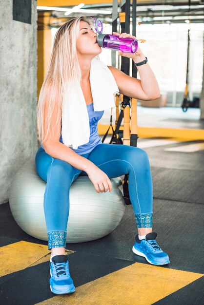 Young woman sitting on fitness ball drinking water in gym