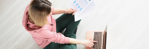 Young woman sitting on floor with documents in her hands and working at laptop top view remote