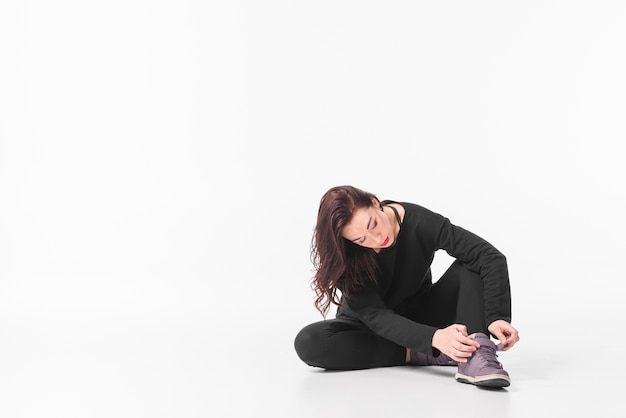Young woman sitting on ground putting shoe strap against white background