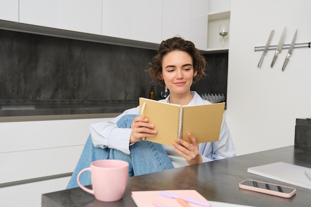 Free photo young woman sitting in kitchen reading her notes in notebook smiling girl at home doing homework