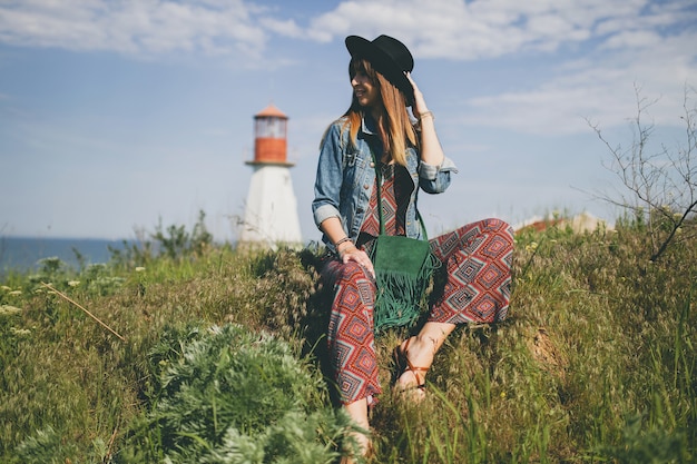 Young woman sitting in nature, lighthouse, bohemian outfit, denim jacket, black hat, smiling, happy, summer, stylish accessories