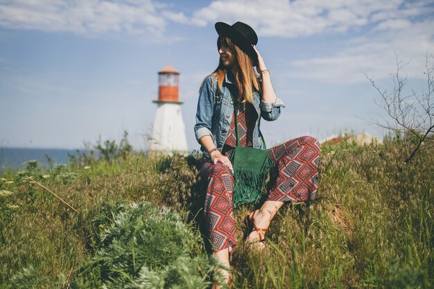Молодая женщина, сидящая на природе, маяк, богемный наряд, джинсовая куртка, черная шляпа, улыбается, счастливая, лето, стильные аксессуары