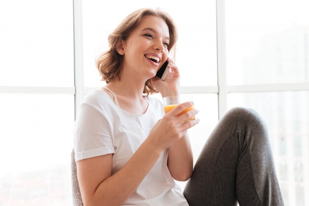 Young woman sitting near window drinking juice talking by phone