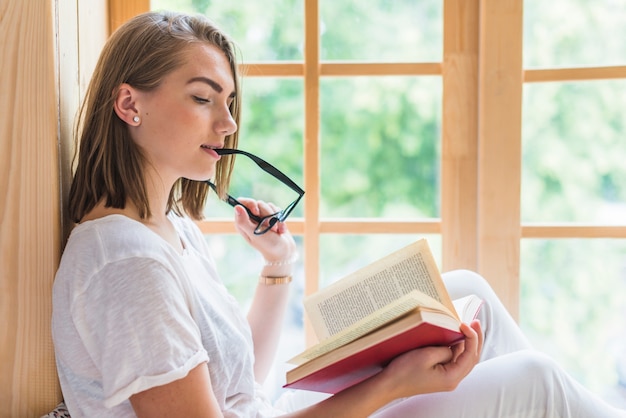 Young woman sitting near the window putting eyeglasses in mouth reading book