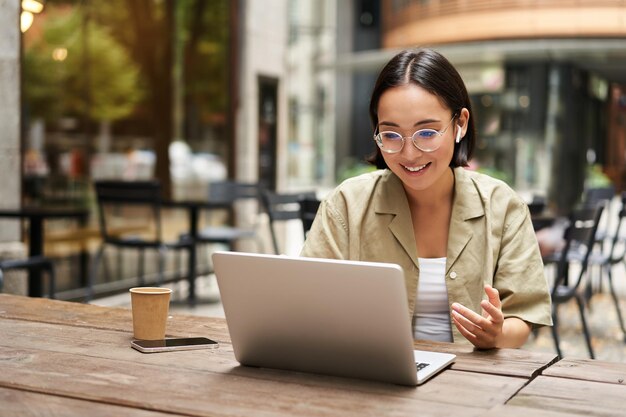 Young woman sitting on online meeting in outdoor cafe talking to laptop camera explaining something