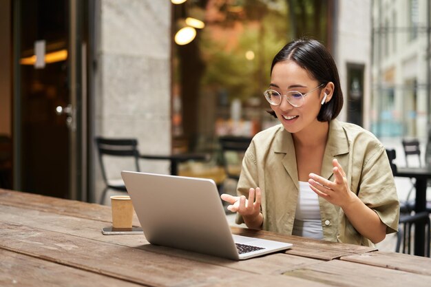 Young woman sitting on online meeting in outdoor cafe talking to laptop camera explaining something