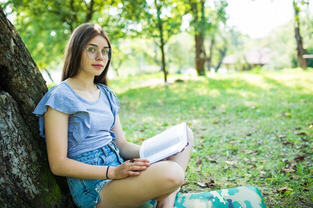 素敵な晴れた夏の木の下でao緑の草に座って彼女のお気に入りの本を読んでいる若い女性