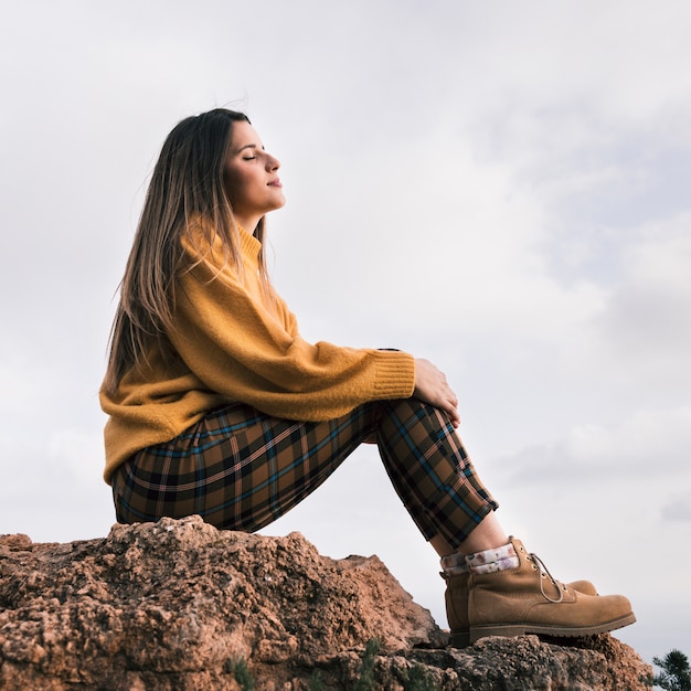 空に対して自然を楽しんでいる岩の上に座っている若い女性