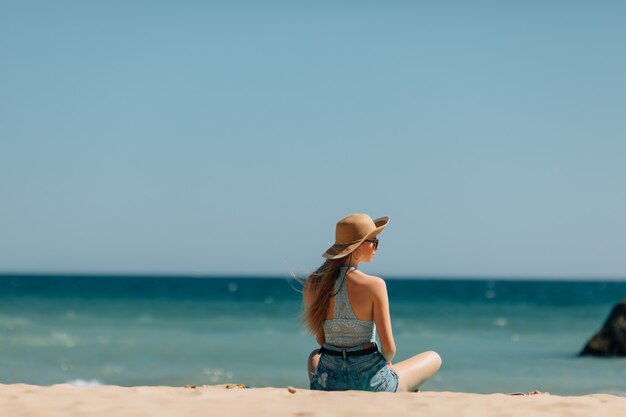 砂の上に座って海を探している若い女性。背面図