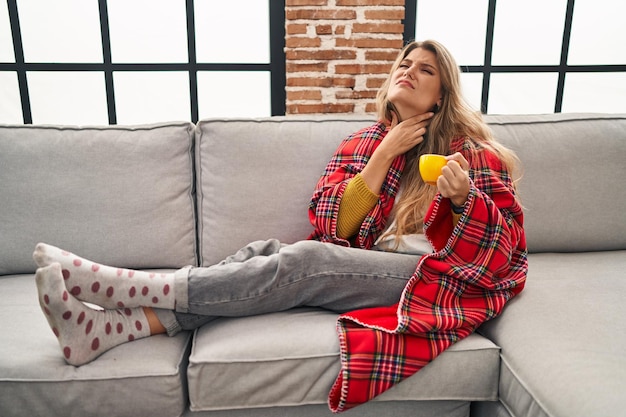 Young woman sitting on the sofa drinking a coffee at home touching painful neck sore throat for flu clod and infection