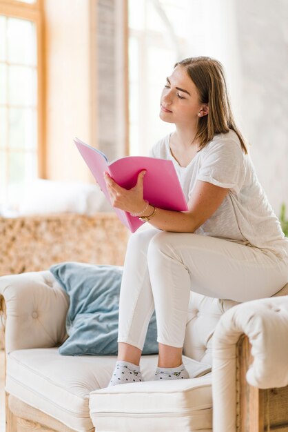 young woman sitting on sofa reading pink book at home