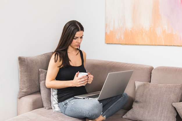 Young woman sitting on sofa using laptop holding coffee cup in hand