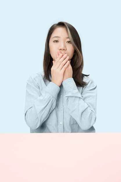 Young woman sitting at table covering her mouth isolated on blue.