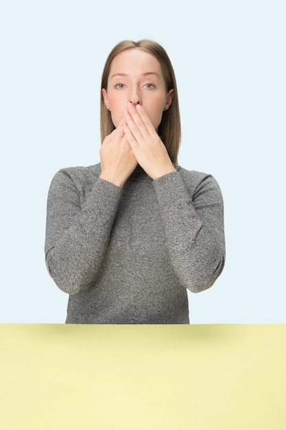 Free photo young woman sitting at table covering her mouth isolated on blue