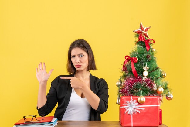 Young woman sitting at a table and pointing her hand in suit near decorated Christmas tree at office on yellow 