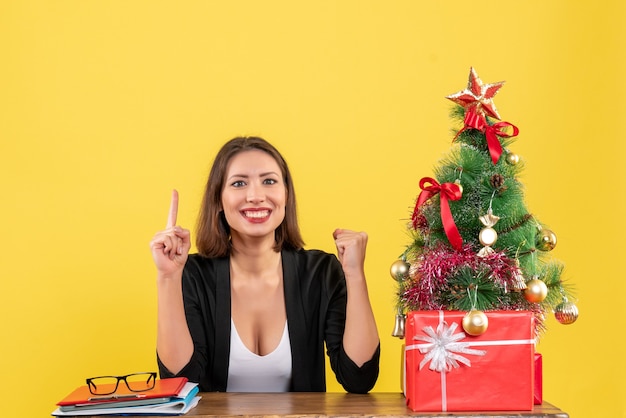 Young woman sitting at a table and pointing up proudly in suit near decorated Christmas tree at office on yellow 