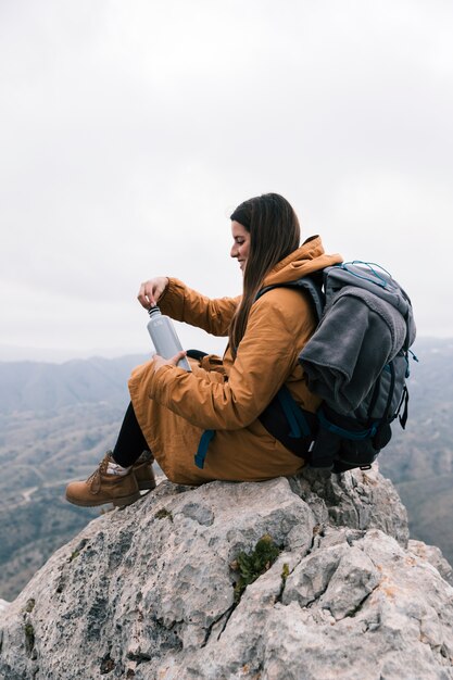 Young woman sitting on the top of mountain with her backpack holding the bottle of water