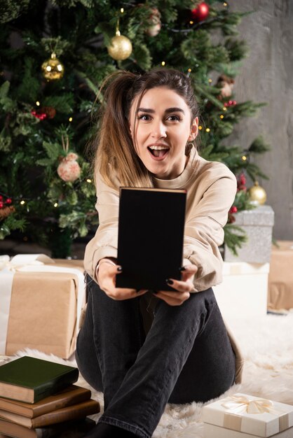 Young woman sitting with black notebook near Christmas tree.