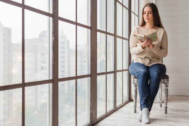 young woman sitting on wooden chair near the window reading the book