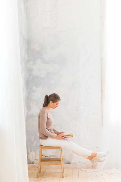 Free photo young woman sitting on wooden stool reading book