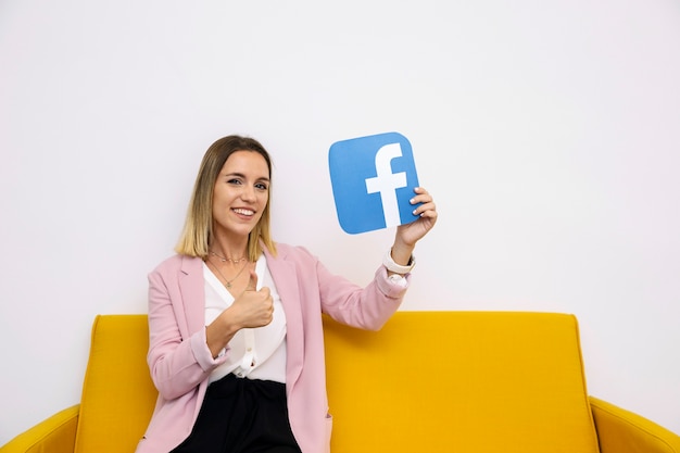 Free photo young woman sitting on yellow sofa holding facebook icon showing thumbup sign