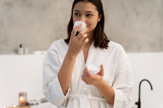 Бесплатное фото Молодая женщина чувствует запах увлажняющего крема после его нанесения на лицо перед принятием ванны