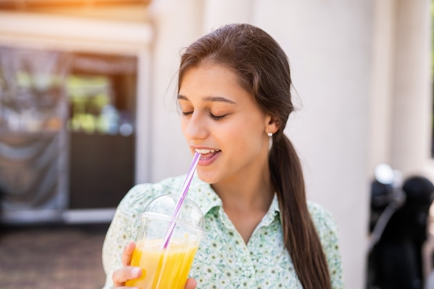 Young woman smiling and drinking cocktail with ice in plastic cup with straw on city street.