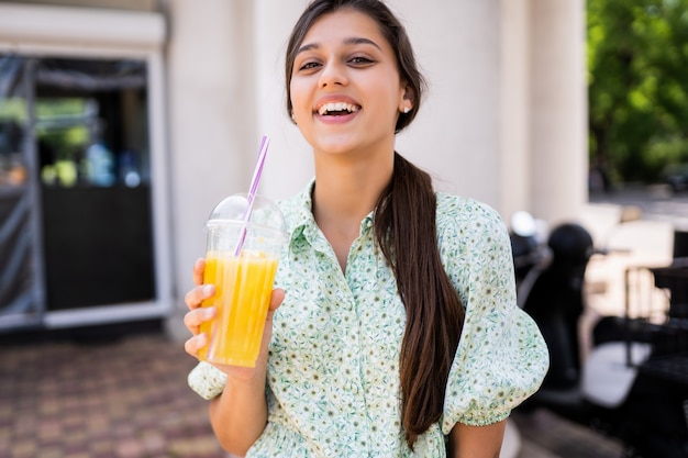 Молодая женщина улыбается и пьет коктейль со льдом в пластиковом стаканчике с соломой на улице города.