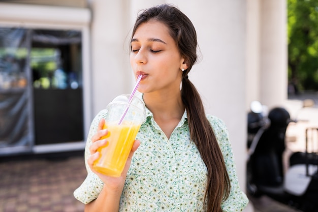 Young woman smiling and drinking cocktail with ice in plastic cup with straw on city street.