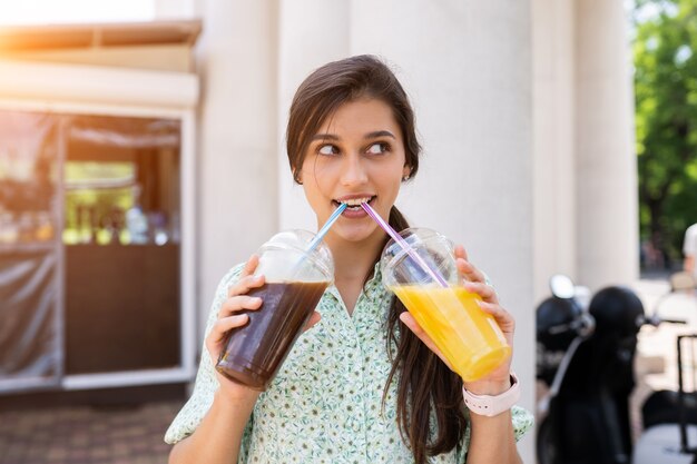 Молодая женщина улыбается и пьет два коктейля со льдом в пластиковых стаканчиках с соломой на улице города.