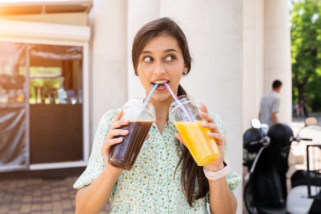 Free photo young woman smiling and drinks two cocktails with ice in plastic cups with straw on city street.