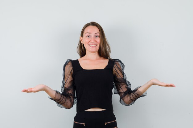 Young woman smiling while spreading her open palms in black blouse and looking glad. front view.